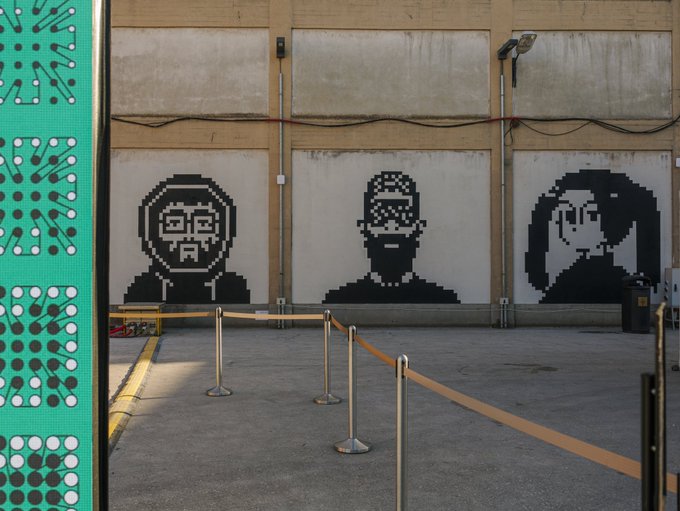 Three large pixel-art style black stencil portraits of figures wearing masks/glasses painted on the panels of a large industrial building, with stanchions and ropes in the foreground.