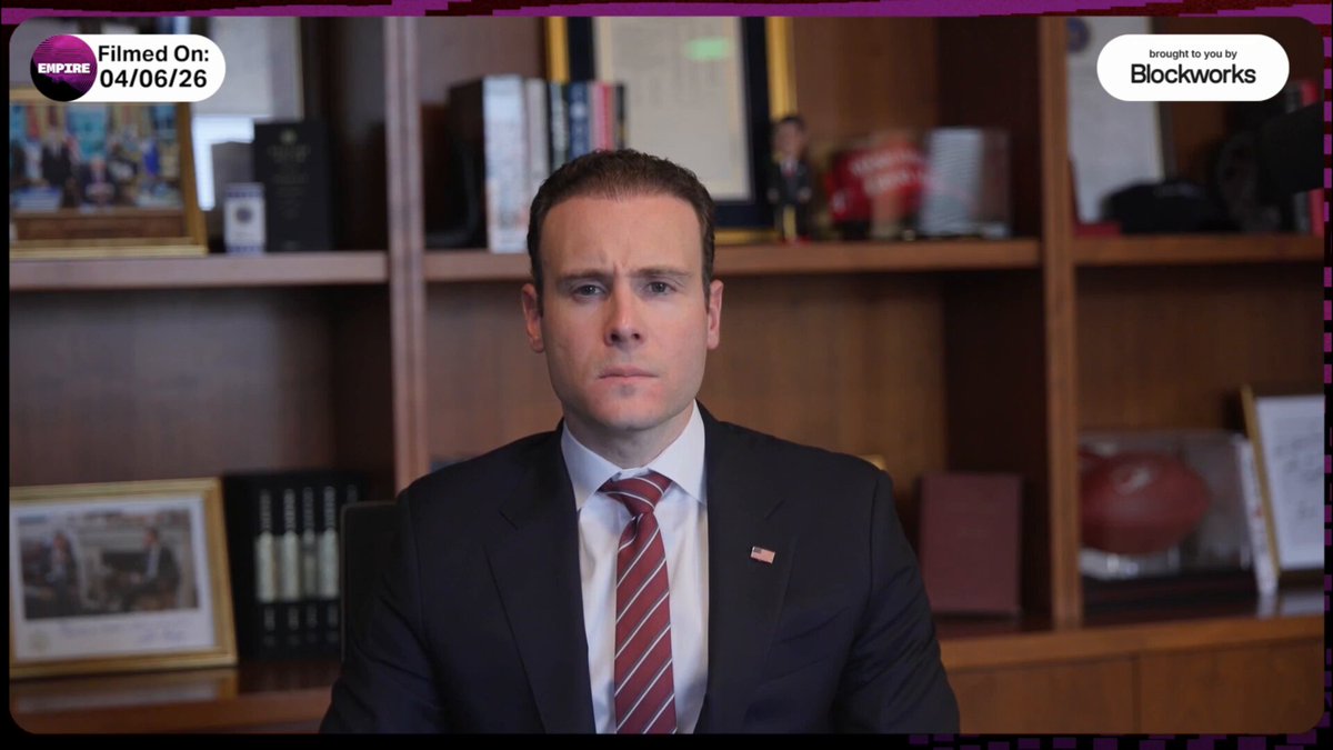 The image shows a man wearing a suit, tie, and jacket sitting at a desk with a computer monitor in front of him. He appears to be looking directly into the camera while speaking or presenting something. The background features a bookshelf filled with various items such as books and figurines. The caption for this image reads: "Hyperliquid".