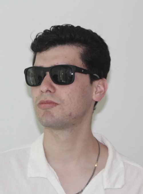 A young man with curly dark hair wears black sunglasses in a Ray-Ban style and a white collared shirt, photographed against a plain background. The image is likely a profile picture or portrait accompanying news about the PumpFun trader known as "Schoen" reportedly outpacing RASMR in trading profits.

No text, numbers, tickers, or names are visible in the image itself.