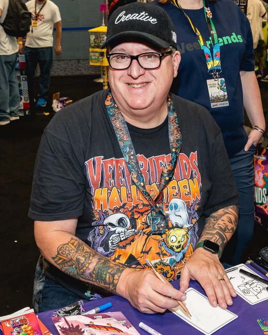 A smiling man wearing glasses and a 'Creative' baseball cap, featuring heavily tattooed arms, signs artwork at a convention table with Halloween-themed merchandise.