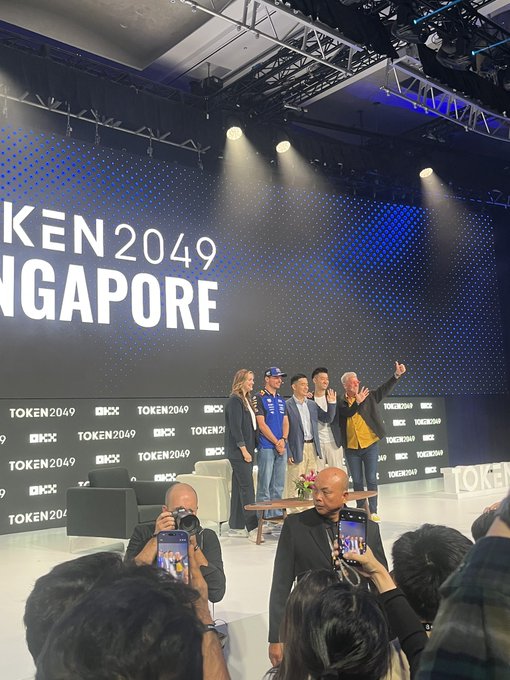 A panel discussion on stage at TOKEN 2049 SINGAPORE with several people posing for the audience, with 'TOKEN2049' and 'OKX' logos visible on the backdrop and photographers in the foreground.