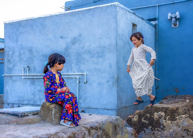 Two young girls interact on a rooftop; one sits on a stone wearing a blue floral dress while the other in a polka dot dress stands nearby.