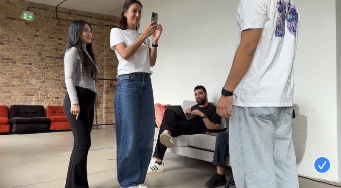 Four people are gathered in a room with exposed brick walls; one woman in a white shirt and jeans stands taking a photo with her phone, another woman stands nearby, a man sits on a sofa, and another man stands in the foreground.