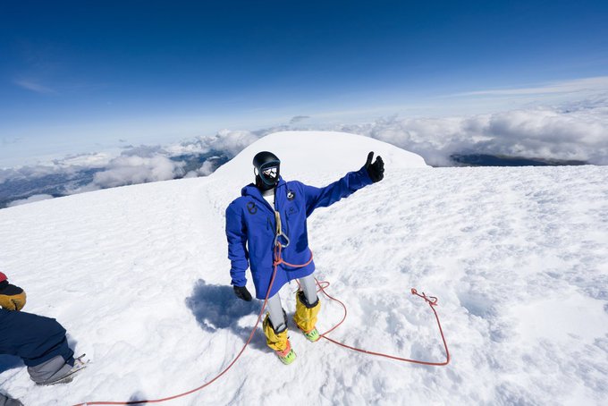 The image shows a person standing on top of a snowy mountain, wearing a blue jacket and yellow boots. The person has their arm raised in the air with a thumbs up gesture. In the background, there are two other people who appear to be observing or participating in the same activity as the main subject. They seem to be at a higher elevation than the main subject, possibly on top of a ski lift or a similar elevated platform. The image also contains text that reads "@OxTrenchor deploy $GOAT and direct fees to @pabloberlangab". This suggests that the person in the foreground might be involved in some form of mountaineering activity or adventure sport, such as skiing or snowboarding, which requires specialized equipment and gear.