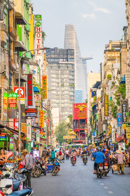 A vibrant street scene in Ho Chi Minh City, Vietnam, featuring motorbikes, colorful signs including 'HOTEL', 'TAN THANH TRADING', and 'NHAT LINH HOTEL', with the Bitexco Financial Tower visible in the background.