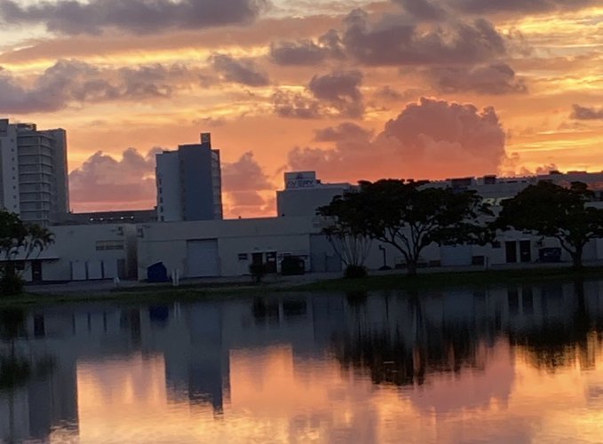 A scenic sunset view with orange and pink clouds reflected in a calm body of water, with buildings and trees silhouetted in the background.