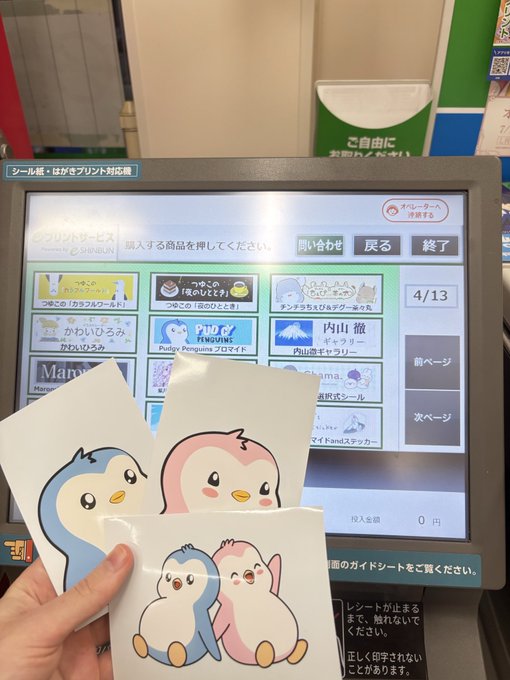 A hand holds three printed stickers featuring cute cartoon characters (blue penguin and pink bear mascots) in front of a Japanese print-on-demand kiosk screen.