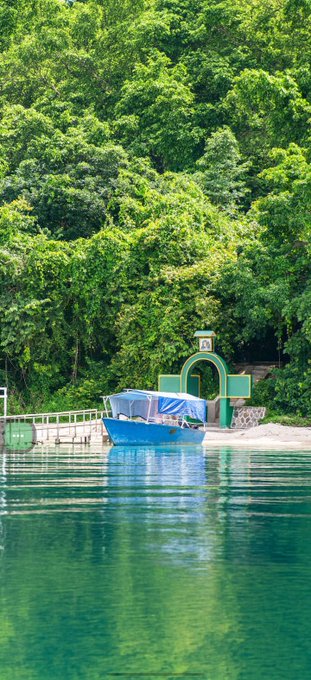 A blue covered boat moored at a small dock with a green archway structure, surrounded by lush green forest and calm turquoise water.