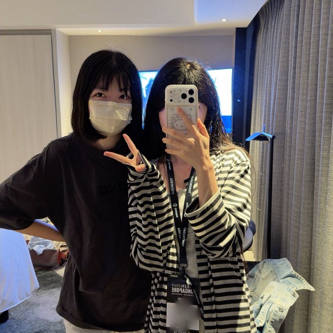 Two young women taking a mirror selfie in a hotel room; one wears a white mask and black t-shirt, while the other in a striped top holds the phone.
