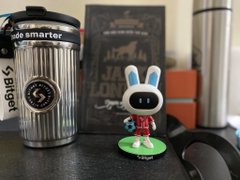 A Bitget-branded metal tumbler with the slogan "trade smarter" sits on a desk next to a small bunny figurine, reflecting the long-awaited arrival of the promotional gift mentioned in the caption. The pairing of the delayed tumbler with the cheerful figurine visually represents the caption's humor about gifts that take so long to arrive that one forgets they even won them.
