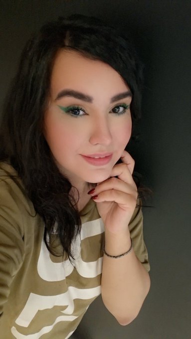 A selfie-style portrait of a young woman with long dark wavy hair, wearing a beige t-shirt and green eyeliner, resting her chin on her hands against a dark background.