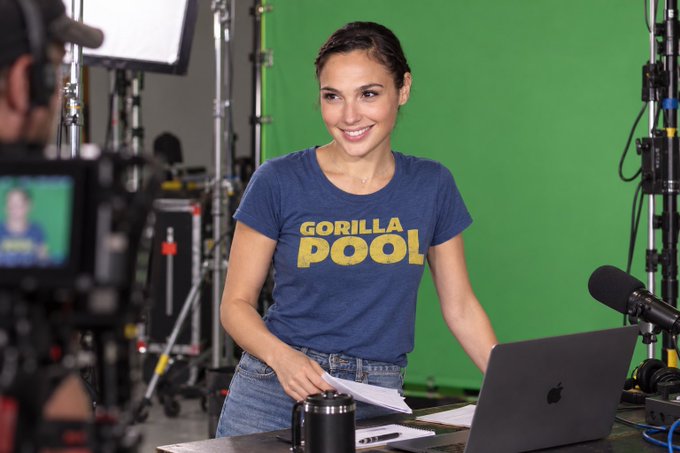 Gal Gadot smiles on a film set with a green screen behind her, wearing a blue 'GORILLA POOL' t-shirt, with a camera, microphone, and MacBook in the foreground.