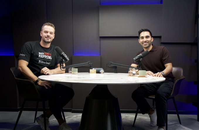 Two men sit across from each other at a round marble table in a studio setting, each with a microphone in front of them, smiling for the camera. The man on the left wears a black "When Sh*t Happens, Bitvise" t-shirt, while the man on the right wears a dark polo shirt, appearing to be Kash Dhanda, COO of Jupiter Exchange, during a podcast recording.