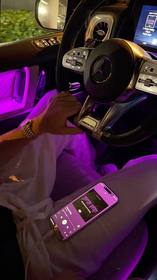 A view from the driver's seat of an AMG Mercedes at night, featuring purple ambient lighting, with the driver holding the steering wheel and a phone playing a song visible on their lap.
