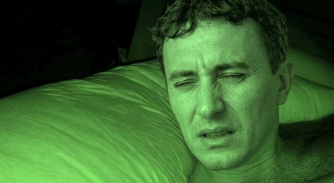 Close-up of a man's face with his eyes closed, illuminated by green night-vision lighting as he lies against a pillow.