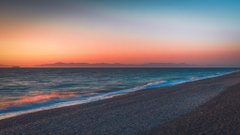 A blurred photograph of a beach at sunset, showing the ocean waves gently rolling onto the shore under a colorful sky with a distant mountain silhouette.