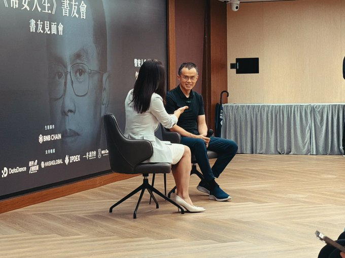 A book talk or panel event with two people seated on stage; a poster behind them shows Chinese text and sponsor logos including BNB Chain, DotDance, HashGlobal, and IPDEX.