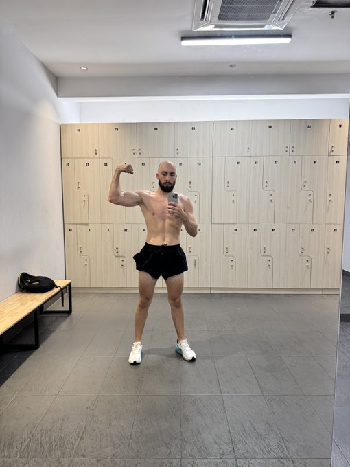A shirtless, muscular bald man with a beard flexes his bicep while taking a mirror selfie in a gym locker room, wearing black shorts and white sneakers.