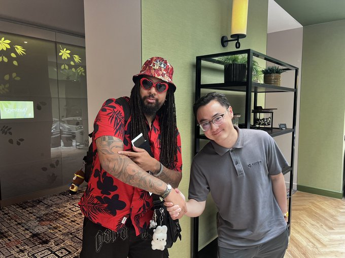 Two men pose for a photo indoors, one with dreadlocks wearing a red Hawaiian shirt and heart-shaped sunglasses shaking hands with another man in a gray polo shirt.