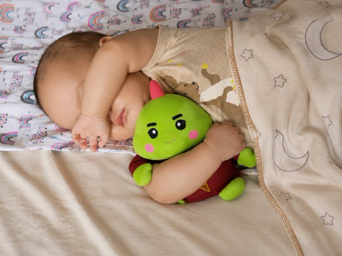 The image features a baby sleeping next to a green stuffed animal, which has a cartoon character design. The scene takes place on a bed with a white comforter and a rainbow-colored blanket. The photo captures the moment of the baby's peaceful slumber, accompanied by the cute green toy that adds a touch of whimsy to the image.