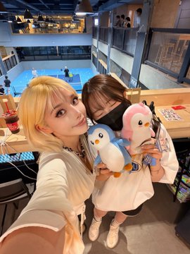 Two young women pose for a selfie indoors, each holding a plush penguin toy, with what appears to be a swimming pool area visible in the background.