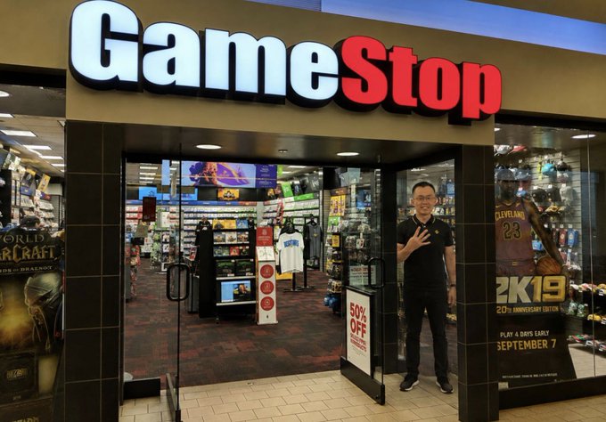 A man poses in the entrance of a GameStop store, making a hand gesture, with a '50% OFF' sign visible and NBA 2K19 and World of Warcraft promotional displays in the windows.
