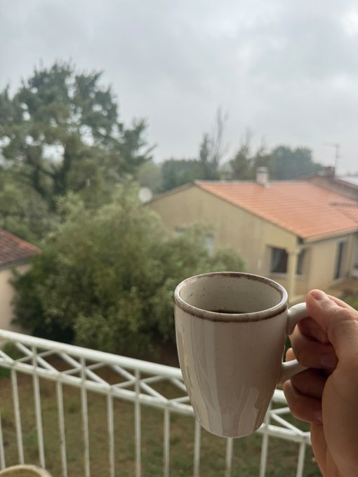 A hand holds a beige ceramic mug filled with dark coffee on a balcony, with a view of a yellow house, trees, and a cloudy sky in the background.