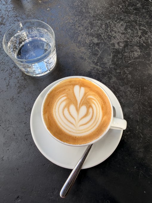 A cappuccino with beautiful latte art sits on a white saucer with a spoon, accompanied by a glass of water on a dark, textured table — the perfect start to a morning with coffee!