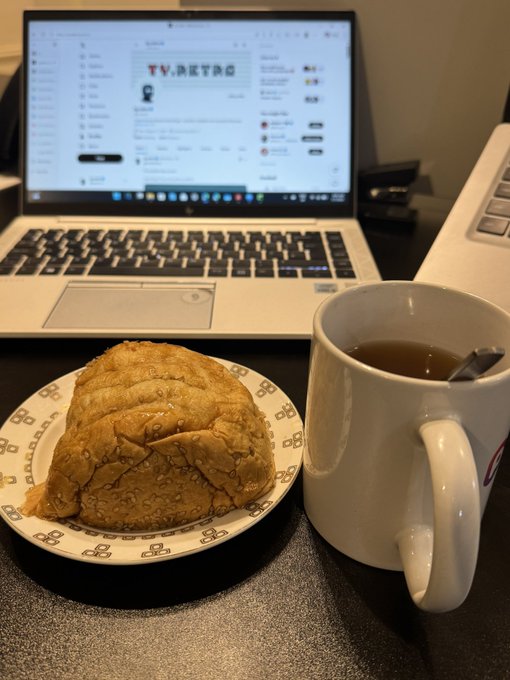 A breakfast or snack setup featuring a sesame-topped pastry on a decorative plate next to a white mug of tea, with a laptop displaying a 'TV.RETRO' webpage in the background.