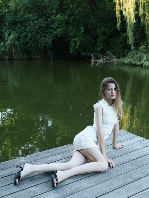 A photograph of a young woman with long blonde hair wearing a white dress and black heels, sitting on a wooden dock by a lake surrounded by green trees.