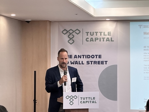 A man in a suit speaks at a podium for Tuttle Capital, with a banner reading "Tuttle Capital" and "The Antidote to Wall Street."