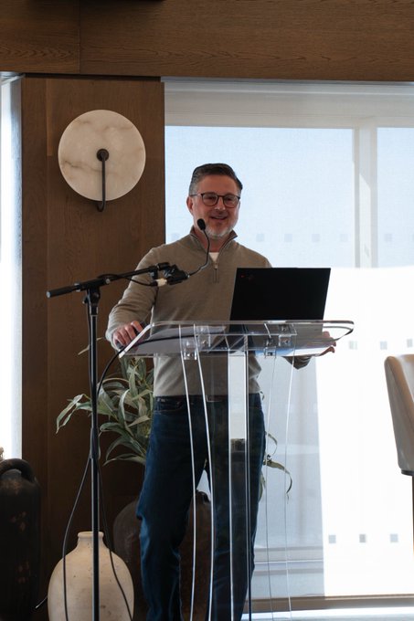 A man stands at a clear acrylic lectern with a laptop and microphone, smiling during a presentation in a modern wood-paneled room.