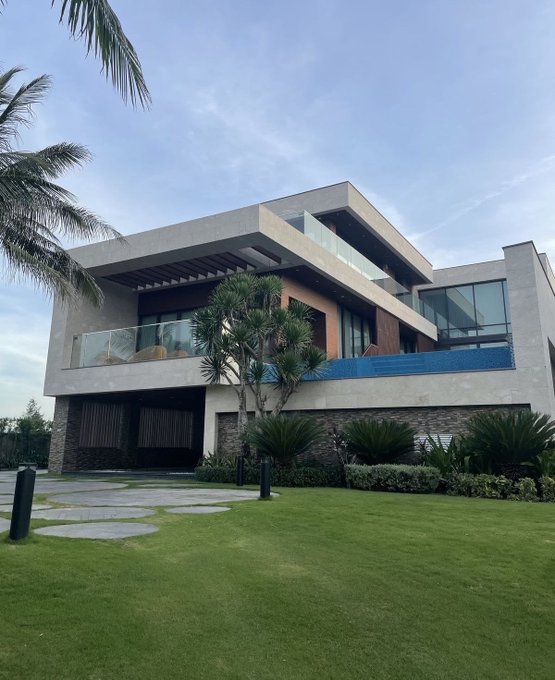 A modern, multi-story luxury home is photographed from the front yard, featuring clean architectural lines, large glass windows, a small pool on an upper terrace, and surrounding tropical landscaping under a clear sky.