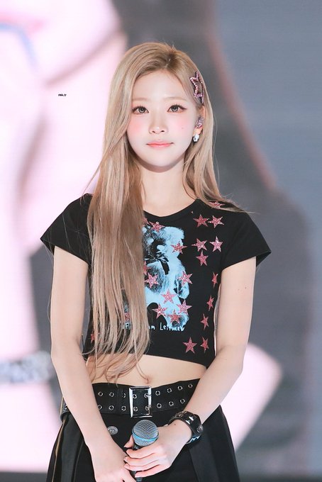 A young woman with long blonde hair holding a microphone, wearing a black t-shirt decorated with pink stars and a graphic print, posing at what appears to be an event.
