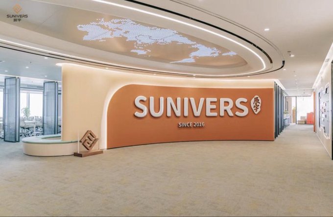 The image shows the modern office lobby of Sunivers, featuring a prominent orange curved wall with "SUNIVERS SINCE 2016" signage, a world map projection on the ceiling, and a small model sculpture on the floor. This corporate setting serves as a backdrop for the post's announcement welcoming a new partner, @tina_nexus, to the "circle," suggesting Sunivers as the workspace where this team collaboration and community membership takes place.