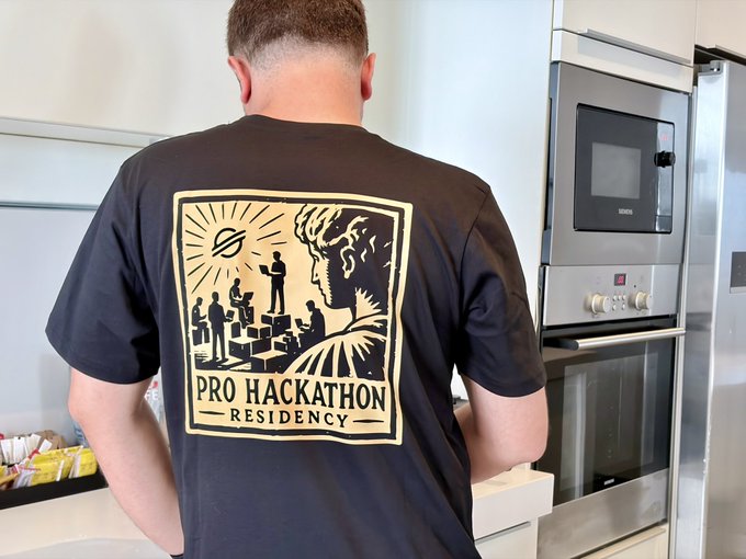 A man wearing a black t-shirt with a graphic reading 'PRO HACKATHON RESIDENCY' stands with his back to the camera in a kitchen.