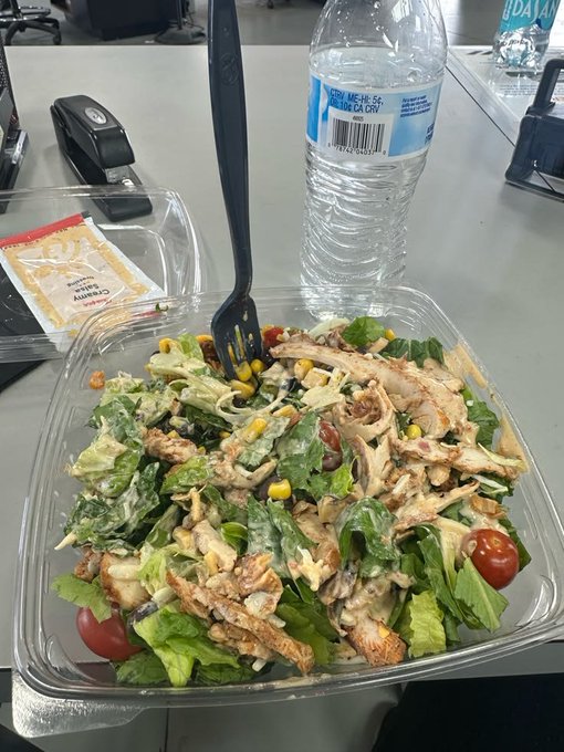 A plastic takeout container holding a salad with grilled chicken, lettuce, cherry tomatoes, corn, croutons, and creamy dressing, with a black fork inserted and a water bottle visible in the background.