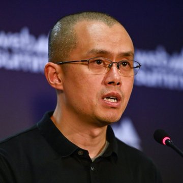 A photograph shows a man with short hair and glasses wearing a black polo shirt, speaking at what appears to be a press conference with a microphone.
