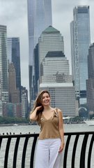 A woman in a tan top and white pants stands at a waterfront railing with the Manhattan skyline, including tall skyscrapers, visible in the background.
