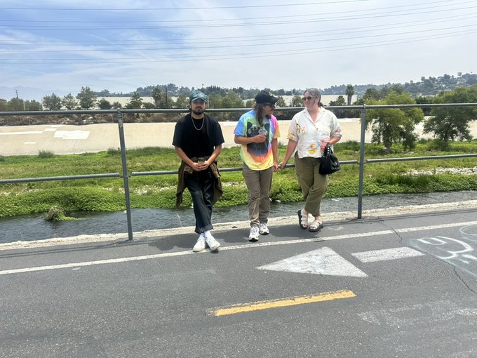 Three people stand on a bike path beside a river, with one man in a tie-dye shirt appearing contemplative, likely after visiting the referenced crypto art exhibition. The post's reflective tone about engaging with @toadswiback's collection aligns with the image's quiet, post-event atmosphere of people processing a meaningful artistic experience together.