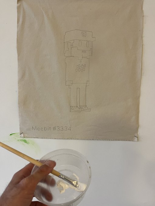A hand-held brush with white paint hovers over a sketch of a blocky character on fabric labeled 'Meebit #3334'.