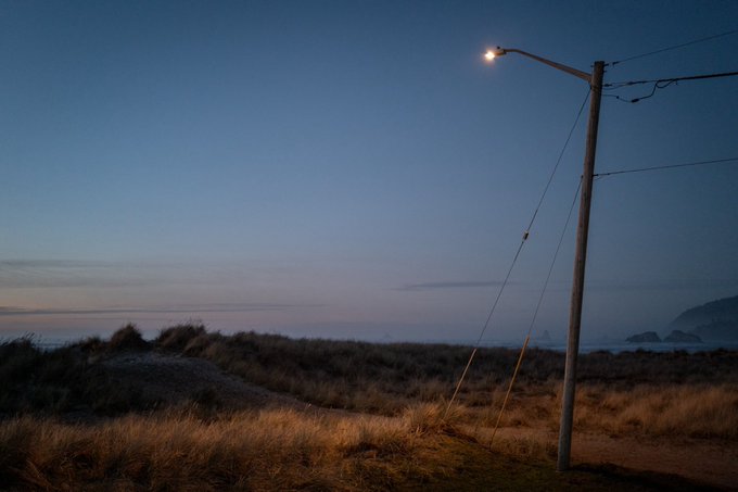 A coastal landscape at dusk or dawn featuring a tall street lamp illuminating dry grass in the foreground, with the ocean and distant cliffs visible under a gradient blue sky.