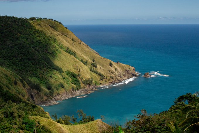 A scenic coastal view shows a green, grassy headland jutting out into a bright blue ocean under a clear sky.