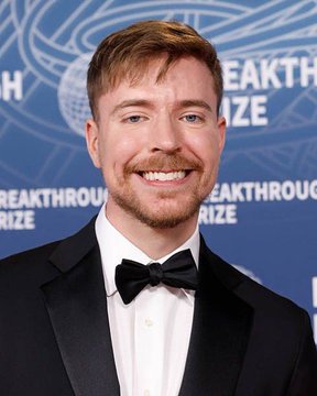 A man, who appears to be MrBeast, smiles at the camera while wearing a black tuxedo and bow tie, standing in front of a blue backdrop that reads "BREAKTHROUGH PRIZE." The image suggests he is attending the prestigious event, potentially setting the stage for his next high-stakes challenge involving 100 crypto traders.