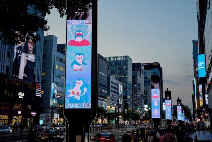 Illuminated vertical billboards display "PENGU x Toys"R"Us advertisements featuring a cartoon penguin in sunglasses across a bustling Seoul street, likely in the Gangnam district, marking the brand's entry into Korean retail.