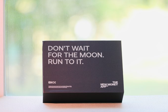 A black desk display with white text reading 'DON'T WAIT FOR THE MOON. RUN TO IT.' alongside 'OKX' and 'THE NEW MONEY APP' branding.