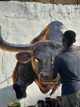 A street artist creating a large mural of a bull on a white brick wall, with paint supplies at the base.