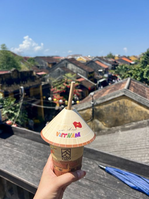 A hand holds a Vietnamese coffee cup from "FaiFo" with a "I love Vietnam" hat topper and a bamboo straw, overlooking a scenic Hoi An rooftops view, contrasting the leisurely "Coffee first, charts later" message with a cultural travel moment rather than the typical Starbucks crypto post.