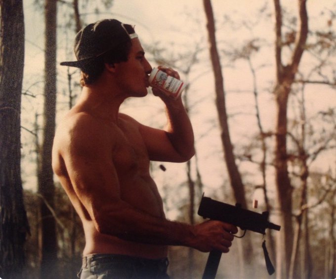 A shirtless young man wearing a backwards cap drinks from a can while holding what appears to be a firearm in a wooded outdoor setting.