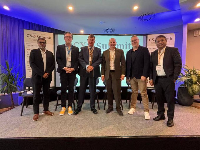 Six men in business attire pose together on a stage at the "CX Summit," with branded banners visible behind them.